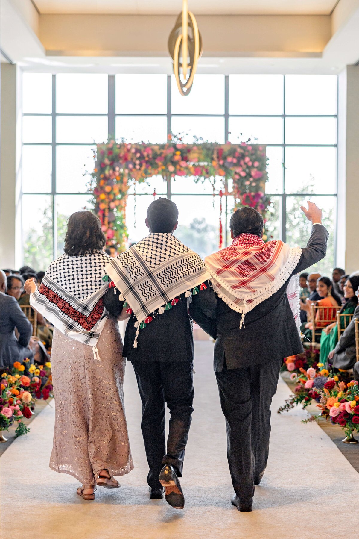 Three people walking down an aisle adorned with colorful flowers, each wearing patterned shawls over their shoulders. They are proceeding towards a floral arch at the front of a room filled with seated guests.