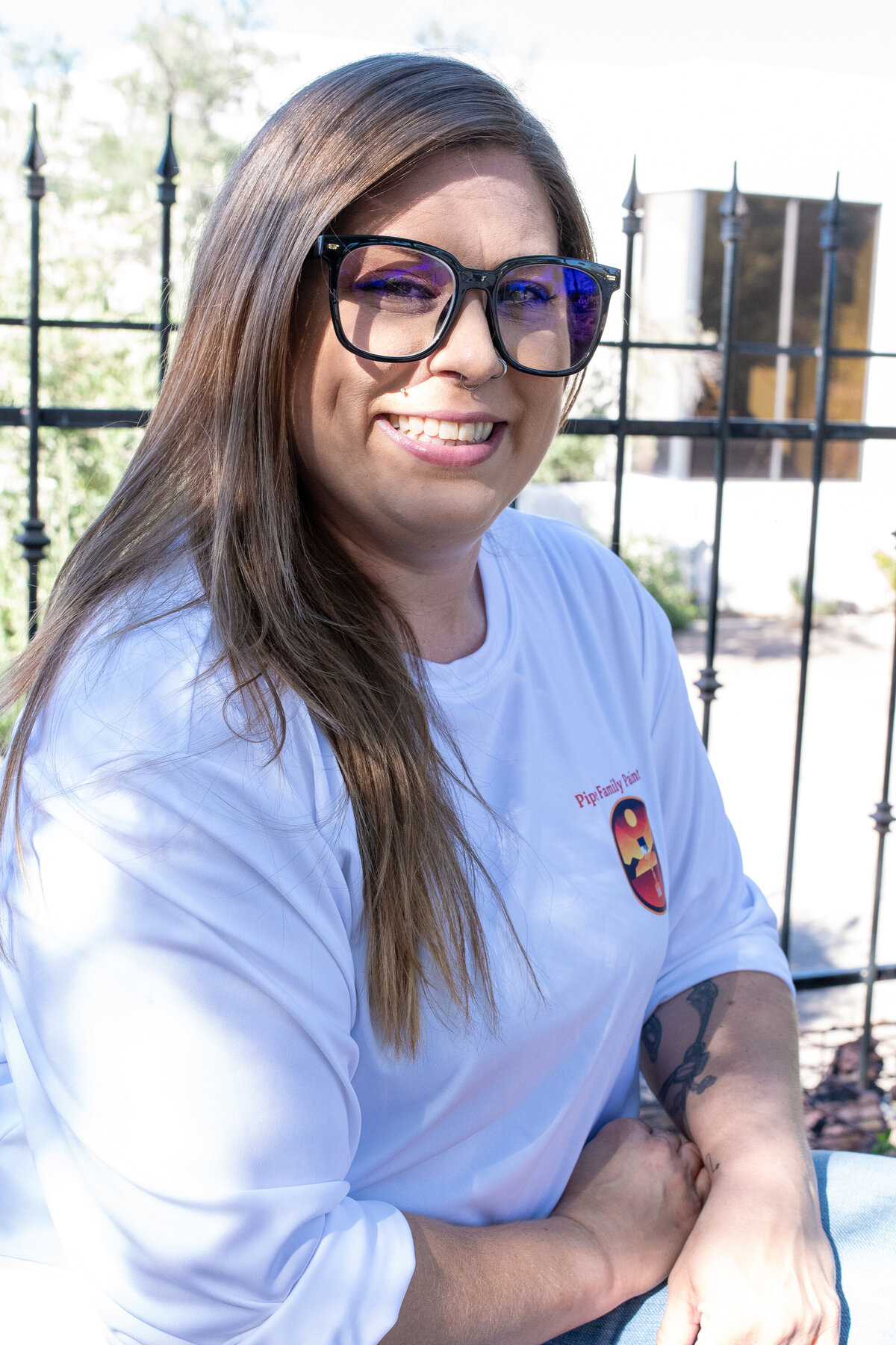 “Smiling woman wearing glasses and a white shirt while seated outdoors near a black iron fence, photographed by Vyrl Photo, showcasing Tucson brand photography.”
