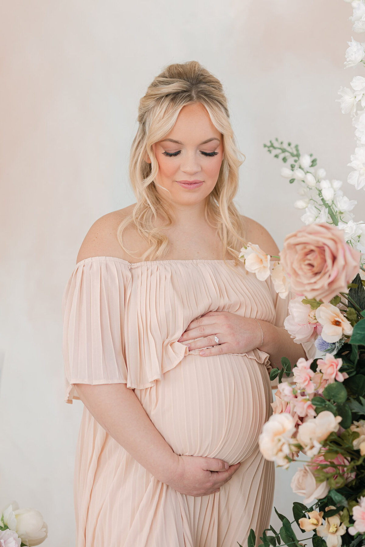 Pregnant mom in pink dress holding her baby bump next to pink flowers in missy marshall photography's louisville kentucky studio