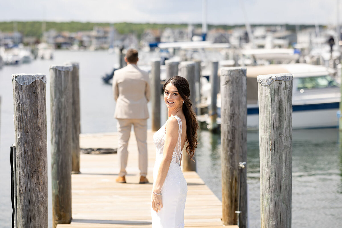 A bride in a white dress smiling over her shoulder on a dock, with a man in a beige suit in the background facing away.