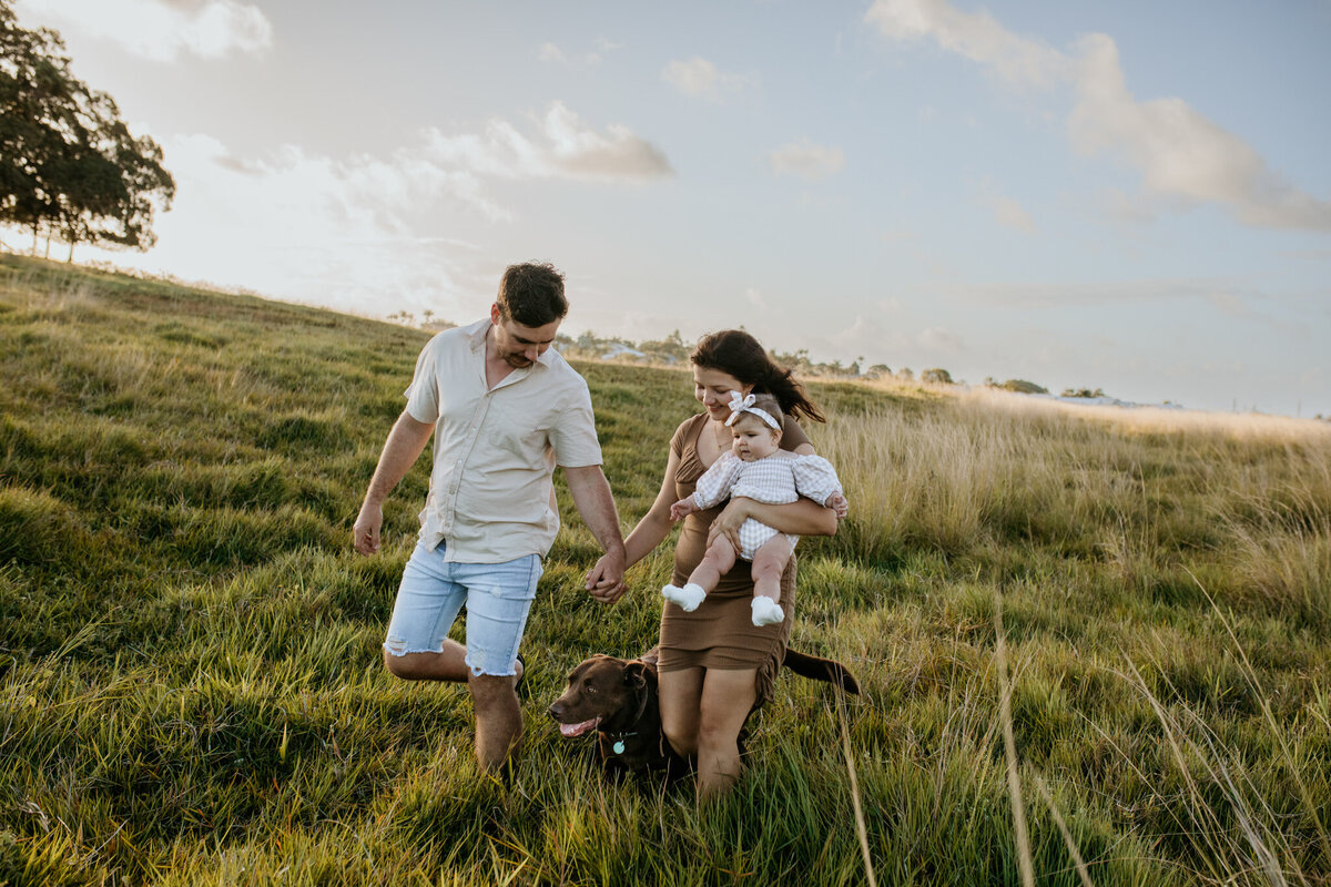 Parents strolling in a field with their dog, mom holding their infant child in her arms.