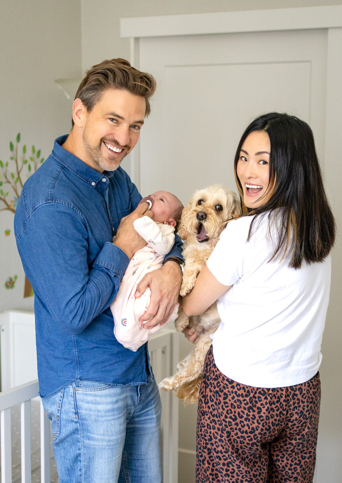 Smiling parents holding their newborn baby and fluffy dog in a bright nursery, captured by San Francisco newborn photographer.