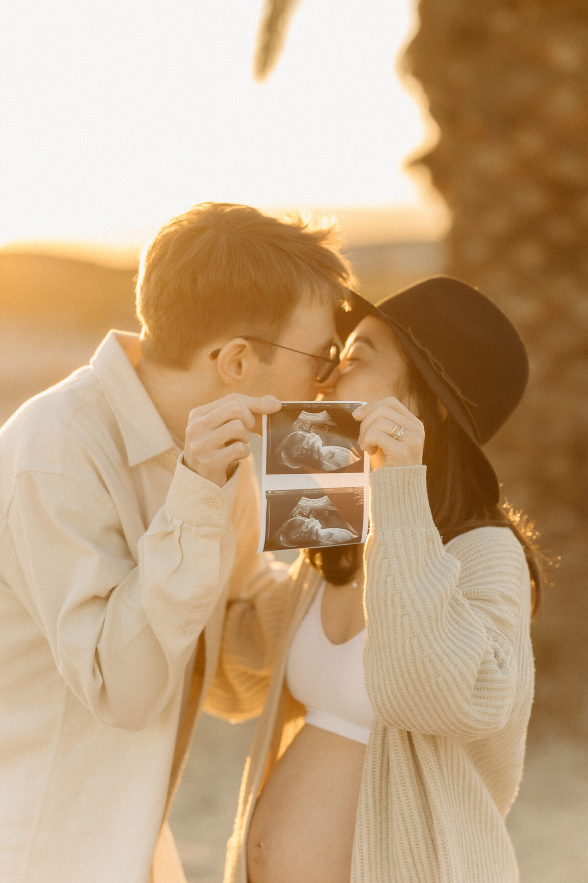 Couple kissing while holding an ultrasound picture at Coronado Beach during golden hour maternity photos. Captured by San Diego Family Photographer Yuka Schad.