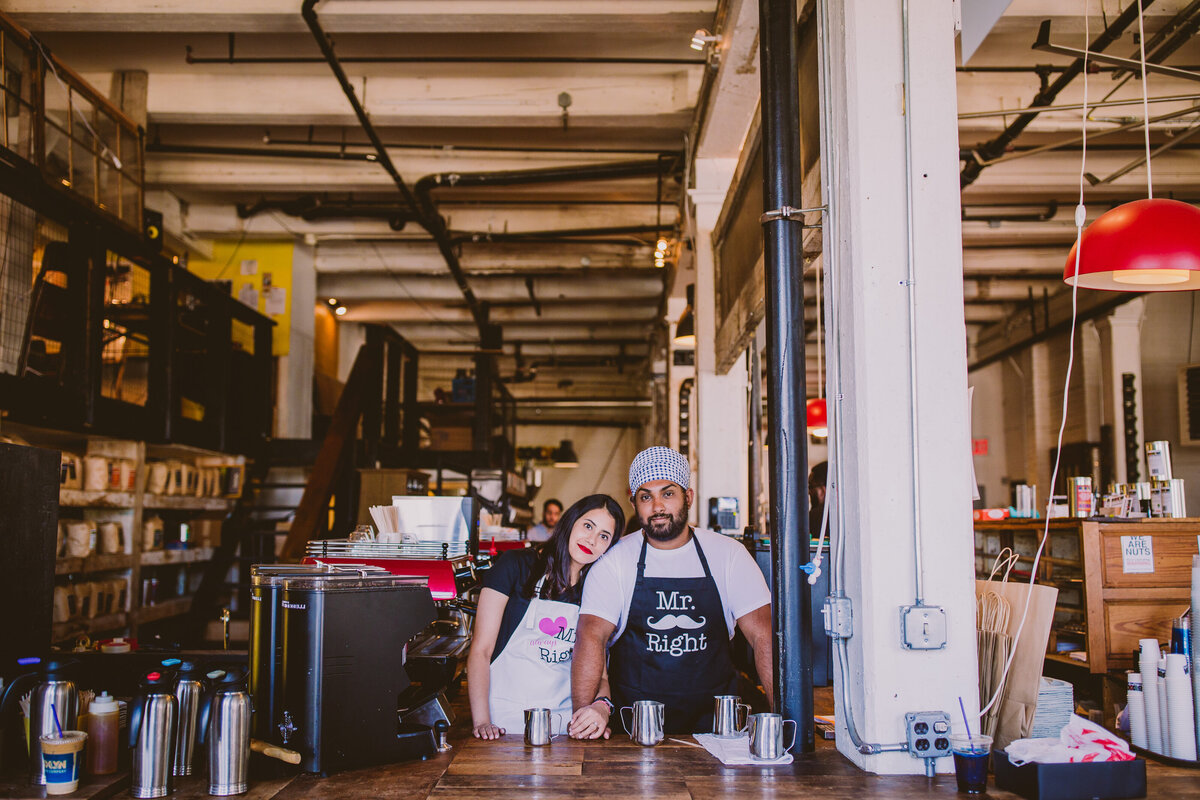 A couple posing behind the counter at the brooklyn coffee roasters in dumbo nyc during their engagement session wearing aprons