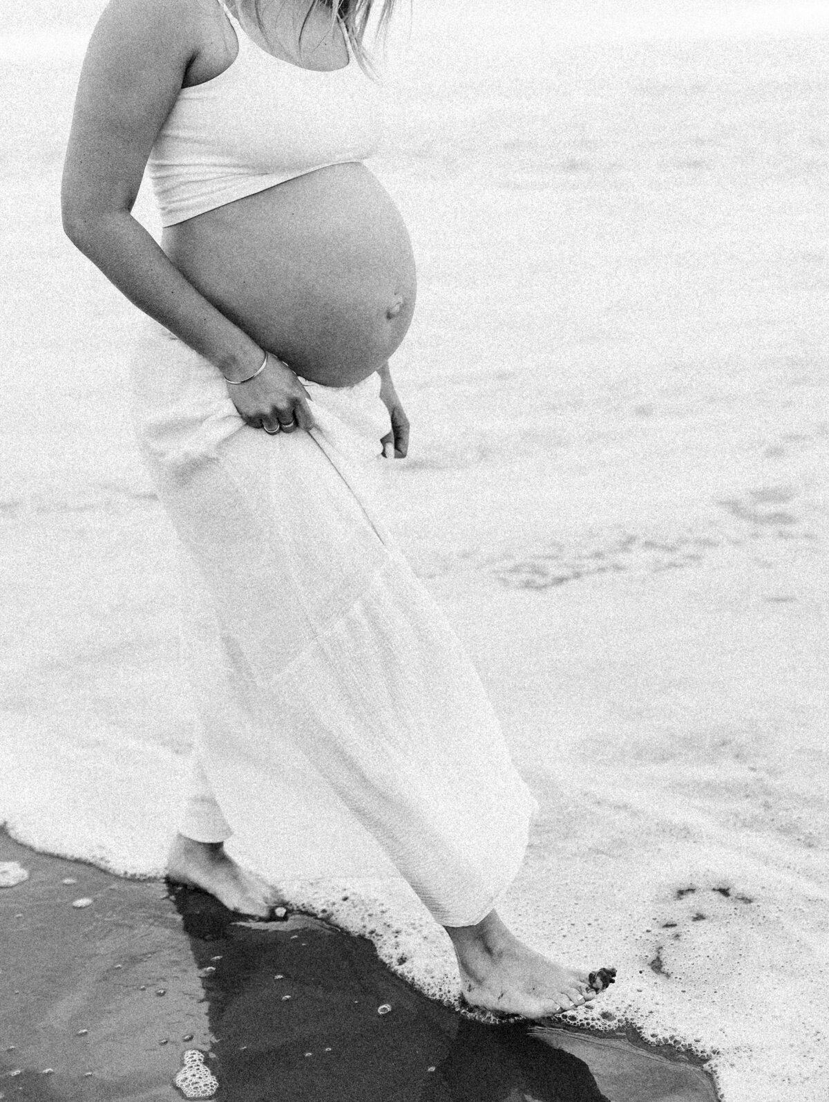 A grainy black and white film image of a pregnant woman walking along a christchurch beach during a maternity photoshoot with lucy jane