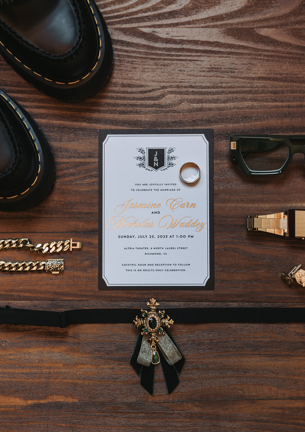 Flatlay detail photo of a wedding invitation suite at the Altria Theater in Richmond, Virginia. The photo includes a wedding invitation, gold ring, black shoes, gold watch, cufflinks, glasses, and accessories arranged on a wooden surface. Shot by Quinn Photography LLC.