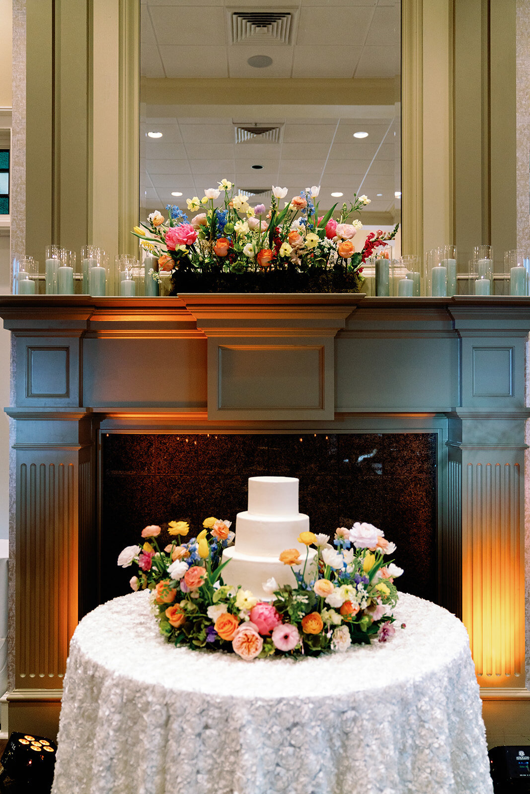 Three-tier white wedding cake surrounded by a vibrant floral ring, displayed in front of a decorated fireplace at Burritt on the Mountain.