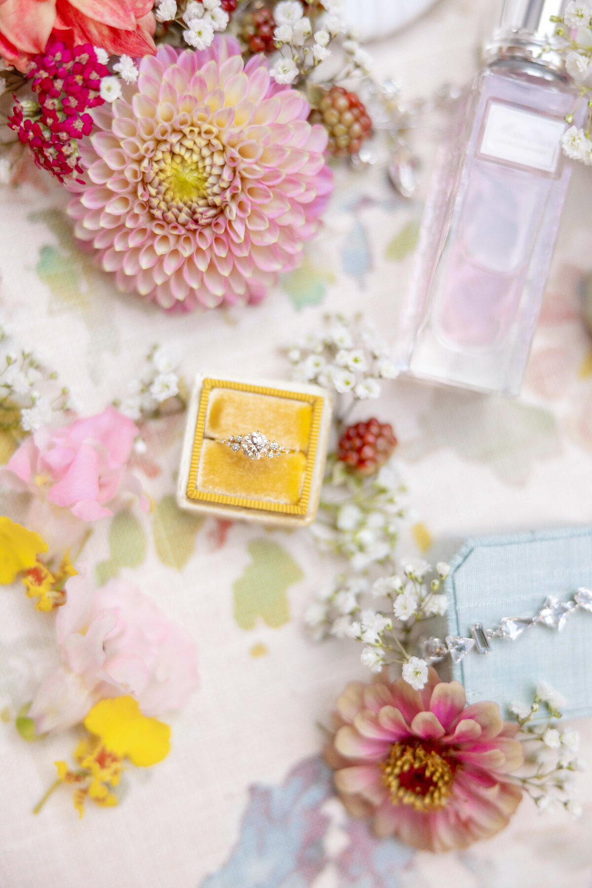 A floral-themed flat lay features a silver ring in a yellow box, surrounded by pink and peach flowers, small white blossoms, and a bottle of perfume on a pastel floral-patterned surface.