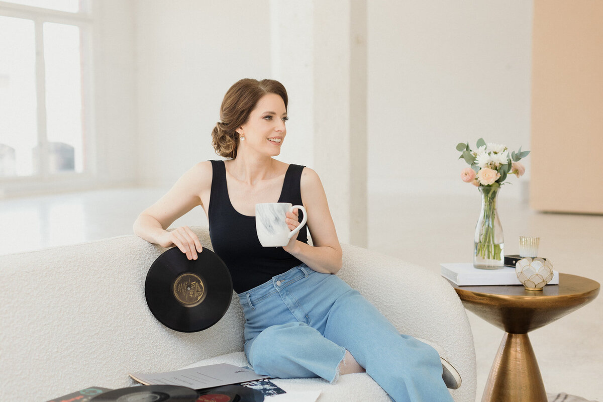 light skinned woman is sitting on a white couch holding tea in one hand an vinyl album in another. More albums are scattered next to her on the couch.