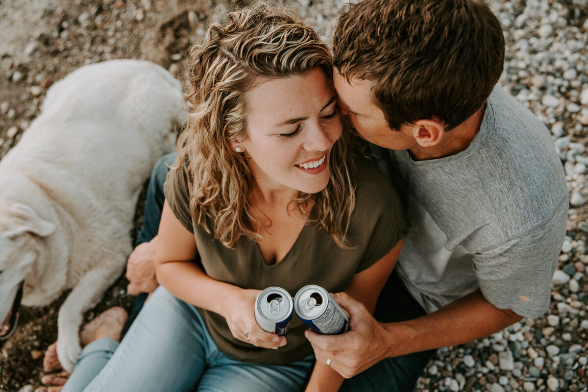 Bride and groom cuddling on stony Mike Weir beach in Sarnia, ON beach holding a beer. The bride has her head resting against the groom's chest and the groom is whispering in her ear. Aerial image.
