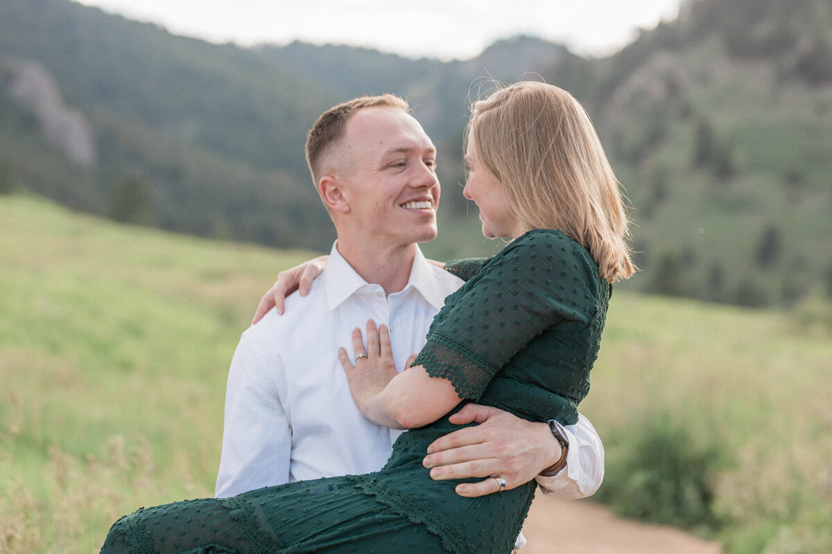 A man beams at his fiancee as he carries her toward the camera, in a meadow overlooking Gregory canyon in Boulder, Colorado
