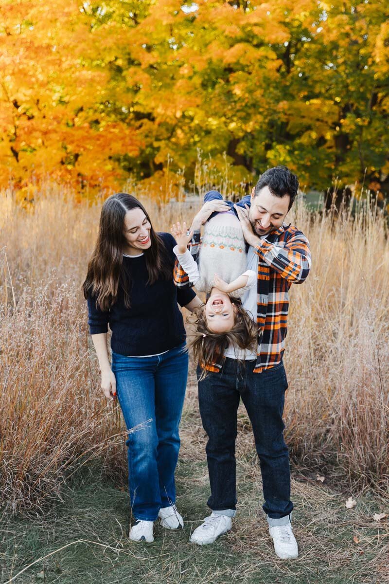 A man playfully holds a child upside down by the legs as a woman stands nearby, smiling brightly. Captured in the vibrant autumn hues of High Park, this joyful moment is expertly framed by a talented family photographer.