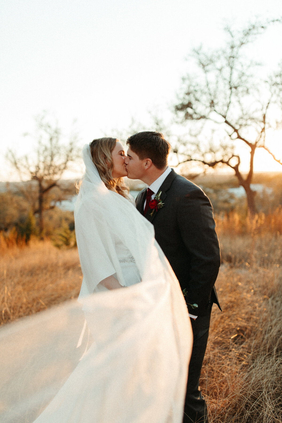 Bride and groom kissing in a field at golden hour while her veil blows in the wind