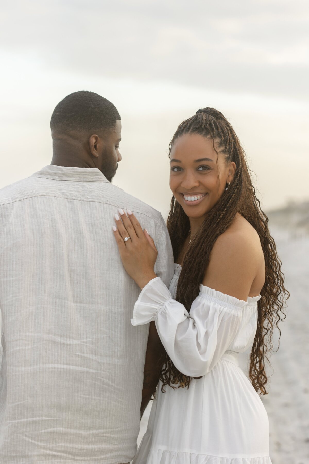A bride-to-be smiles brightly at the camera while resting her hand on her fiancé’s shoulder, showing off her engagement ring during a beach session. Both are dressed in light, neutral outfits, and the soft sandy background is lit by golden sunset light. Captured by Heartful Films, a Jacksonville engagement photographer and videographer known for natural, warm, and joyful storytelling along Florida’s coastline.