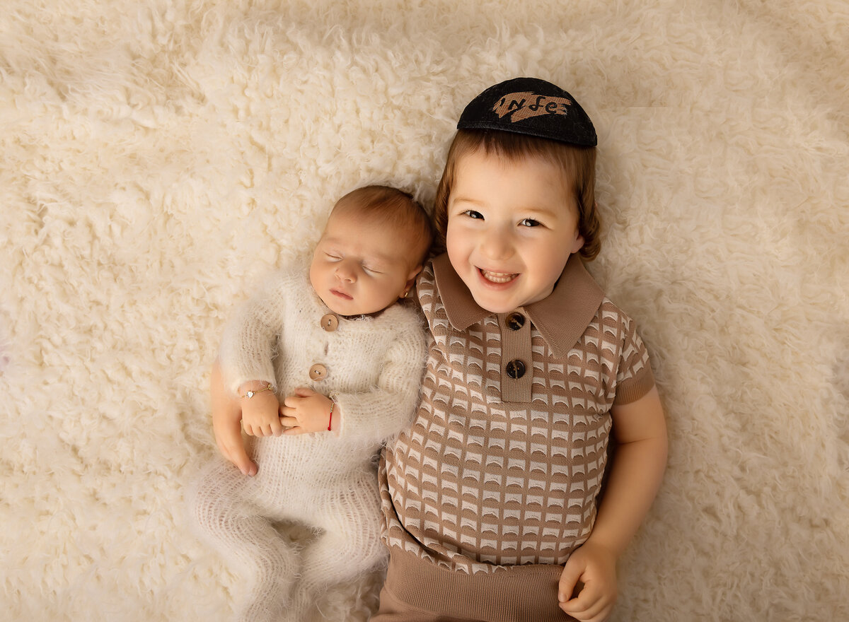 Brooklyn newborn photoshoot. Aerial image. big brother is laing on the ground with his little brother resting on his shoulder. Baby is sleeping and big brother has a big smile at the camera. Captured by top Brooklyn newborn photographer Chaya Bornstein