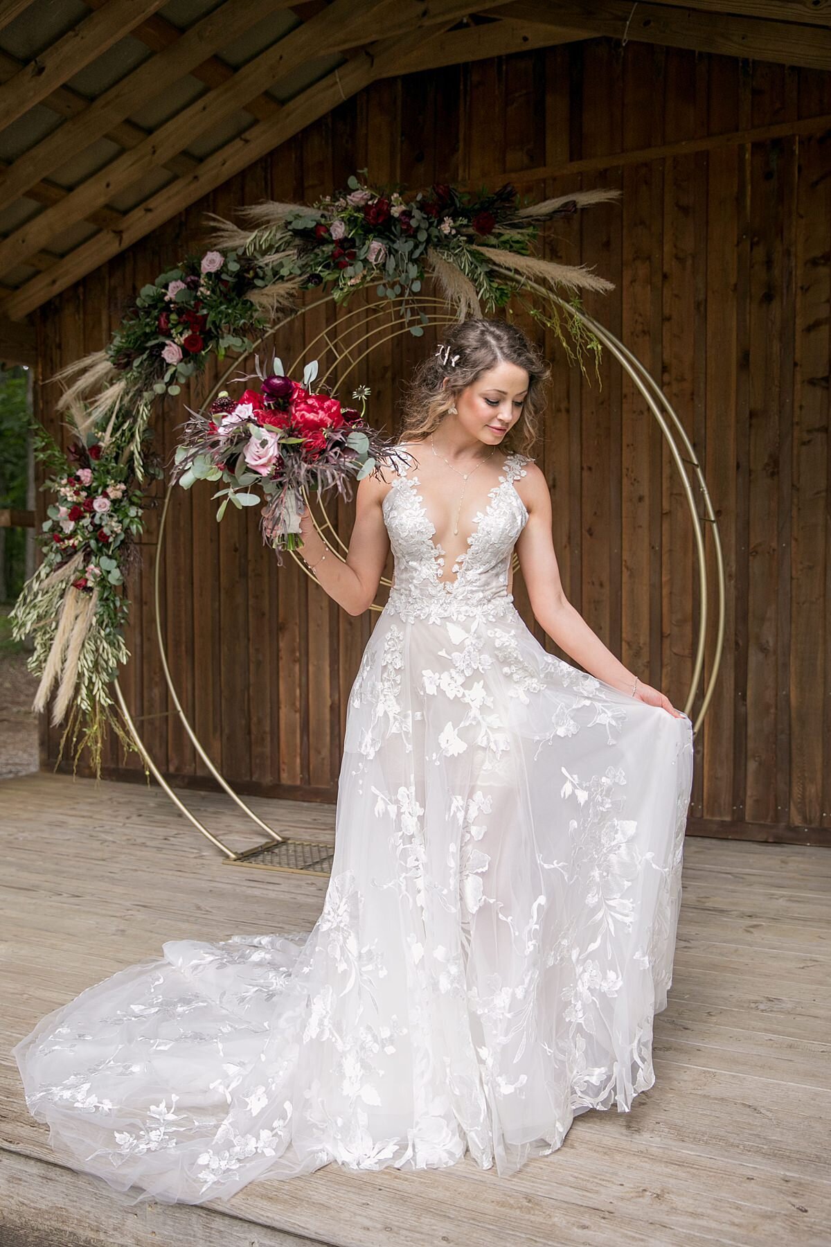 A bride wearing a long lace wedding dress with a deep plunging neckline holding a large bouquet of red peonies, burgundy roses, blush roses and burgundy ranunculus as she stands in front of her round gold metal arbor decorated with pampas grass, red, blush and burgundy flowers as it sits inside a rustic wooded pavilion at Saddle Woods Farm