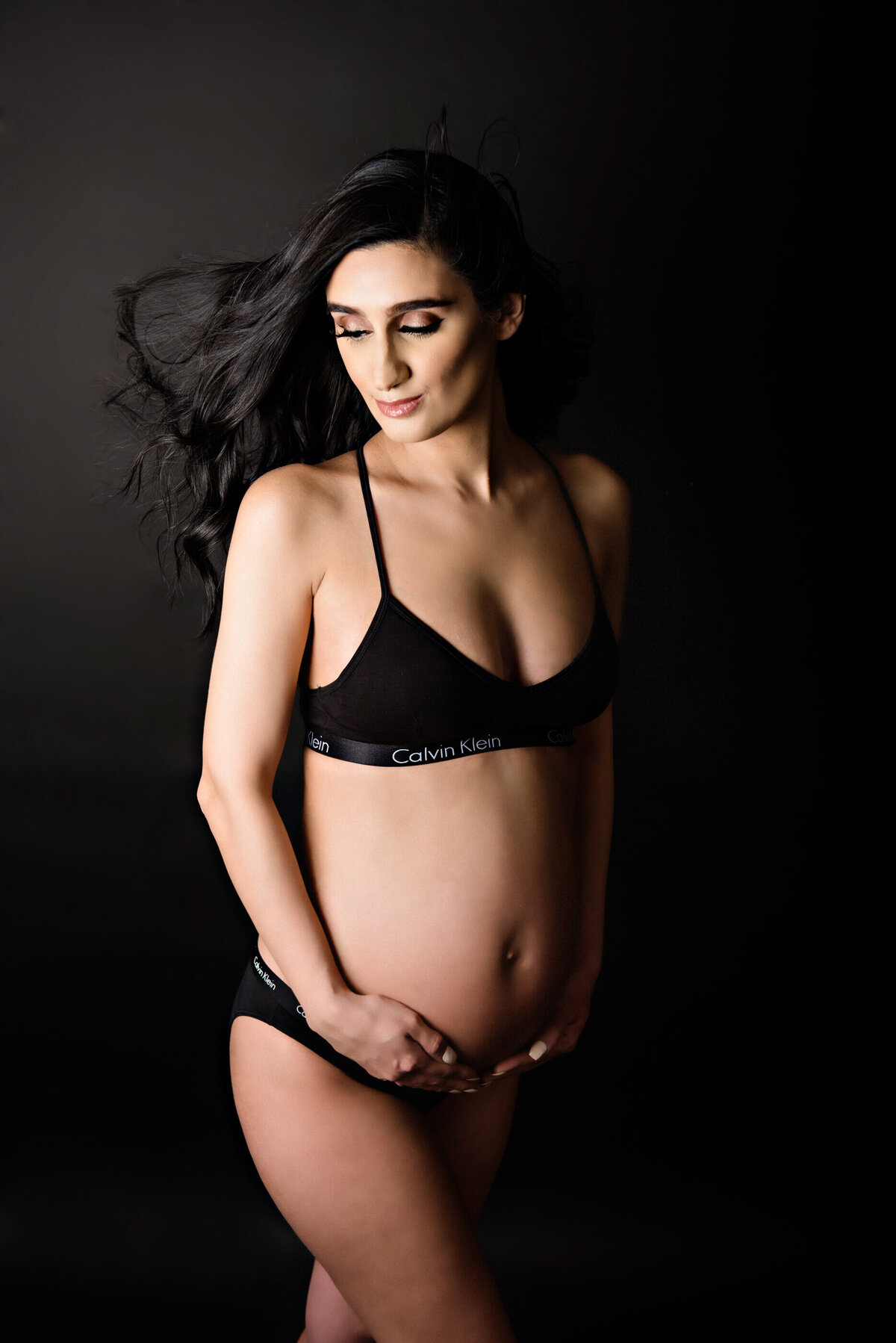 brown-haired pregnant woman with her hair blowing wearing a black bra and underwear for her Burlington studio maternity photography session