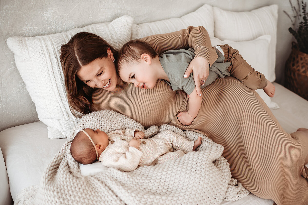 mom snuggled on bed with newborn and son hugging her hip