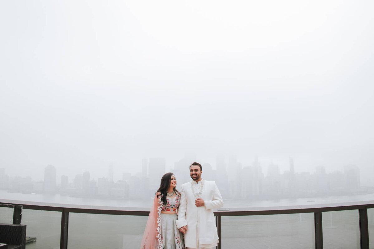 A couple in traditional attire smiles on a foggy rooftop overlooking a city skyline, at Jersey City Hyatt House