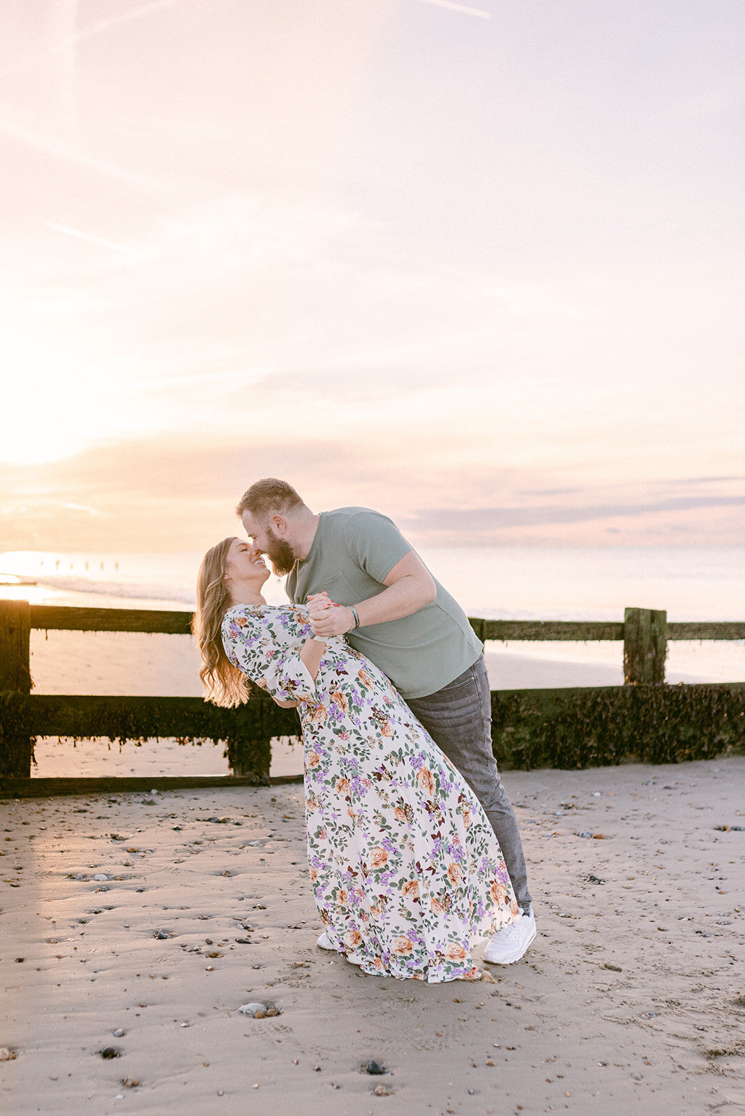 Couple who just got engaged are on the beach, with the sunrising behind them. The lady is wearing a flowery dress and the guy a green tshirt and black jeans, they are holding hands and he is bending her slightly back for a kiss