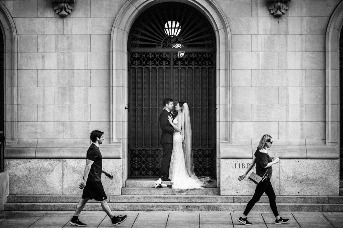 Black and white street scene capturing a bride and groom kissing in front of a library entrance, as passersby walk in the foreground