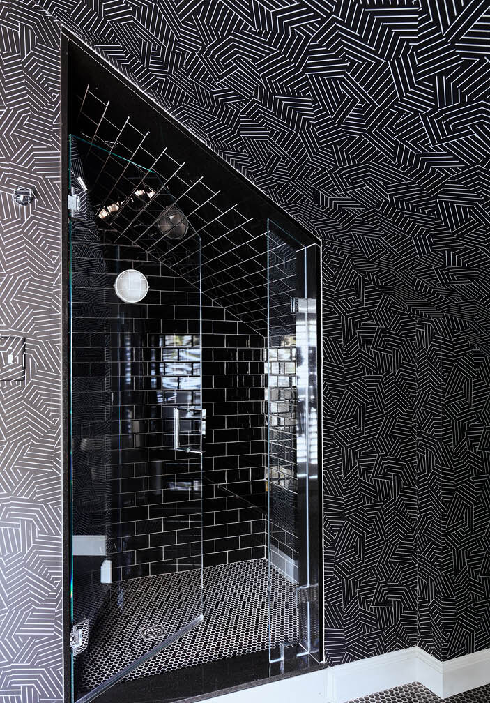 Explore the attic bath in a historic Hingham home, featuring a walk-in shower with black wall and floor tiles, complemented by bold black geometric wallpaper.