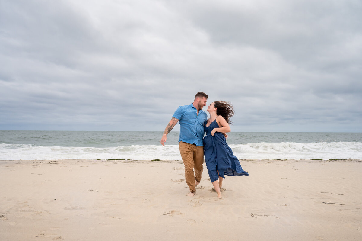 A couple in blue clothing walks hand in hand along a sandy beach with waves in the background under a cloudy sky.