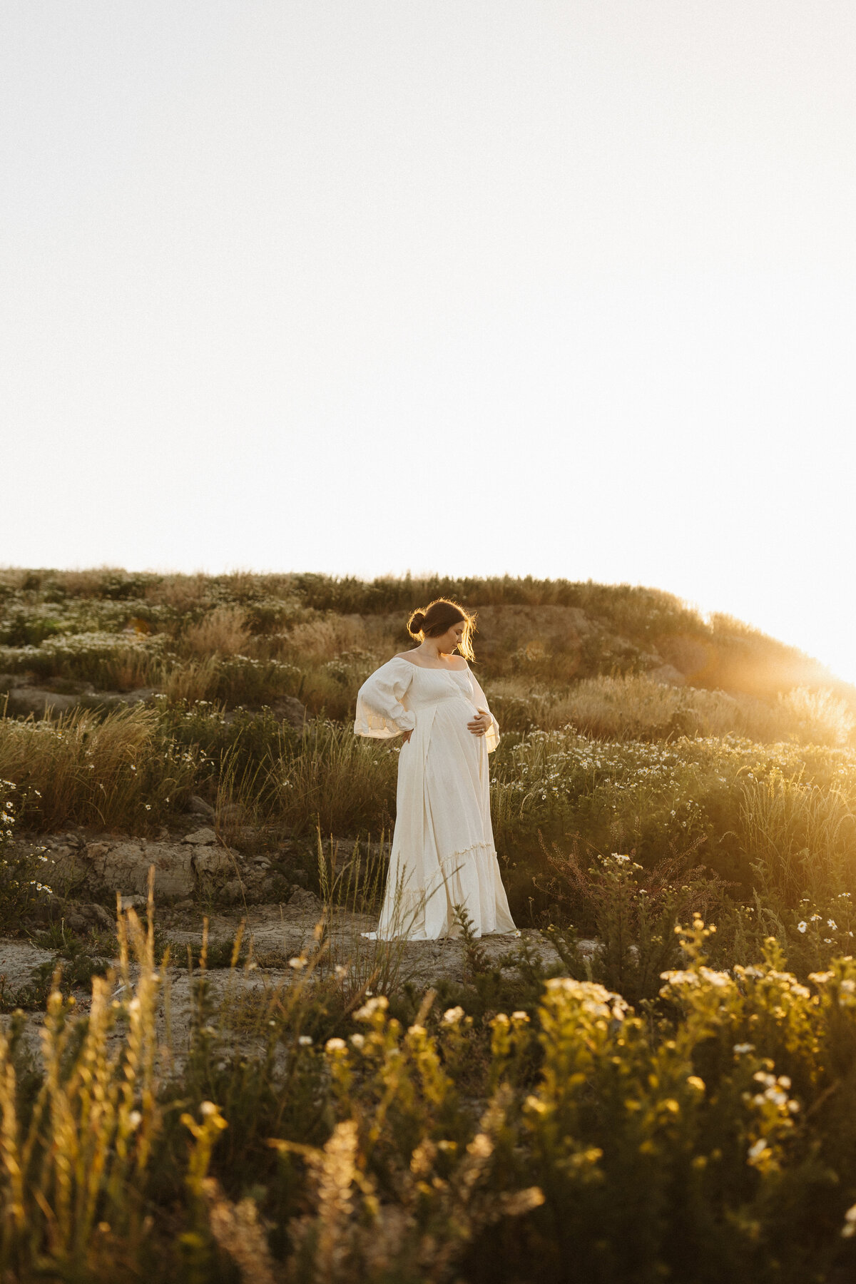 An expective mother to be wears a beautiful dress during a maternity photoshoot and stands in a field of flowers and grasses in christchurch