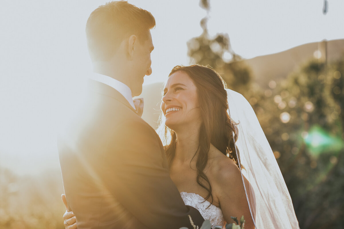 Kohnur Cinematic, romantic couple portrait at golden hour with bride in natural radiant makeup and loose waves, wearing a lace gown and veil on a California hillside.