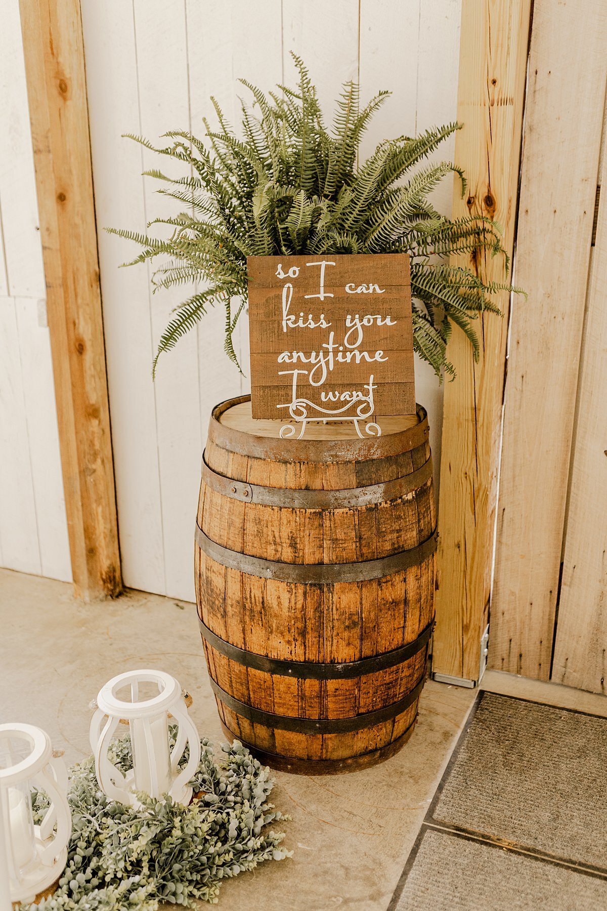 A wine barrel topped with a large green fern inside a white barn had a barn wood sign with a quote from Sweet Home Alabama. Beside the wine barrel are two white lanterns wreathed in greenery.