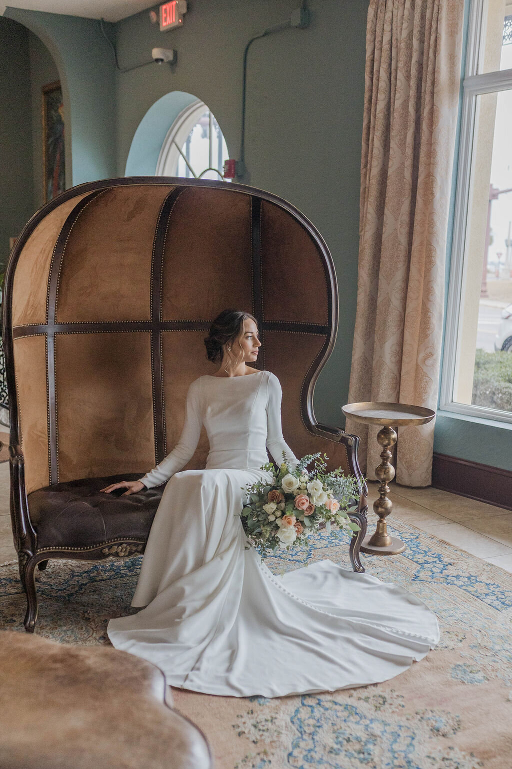 A timeless bridal portrait taken at The White Room in St. Augustine, Florida by Phavy Photography. The bride, dressed in a sleek long-sleeve gown, sits gracefully on a vintage dome-back chair, holding a romantic bouquet with soft pastel florals. Natural light pours in through tall windows, highlighting the elegant textures and soft tones of the space. Captured in a true-to-color, documentary style that reflects Phavy Photography’s signature approach—authentic, editorial, and full of emotion.