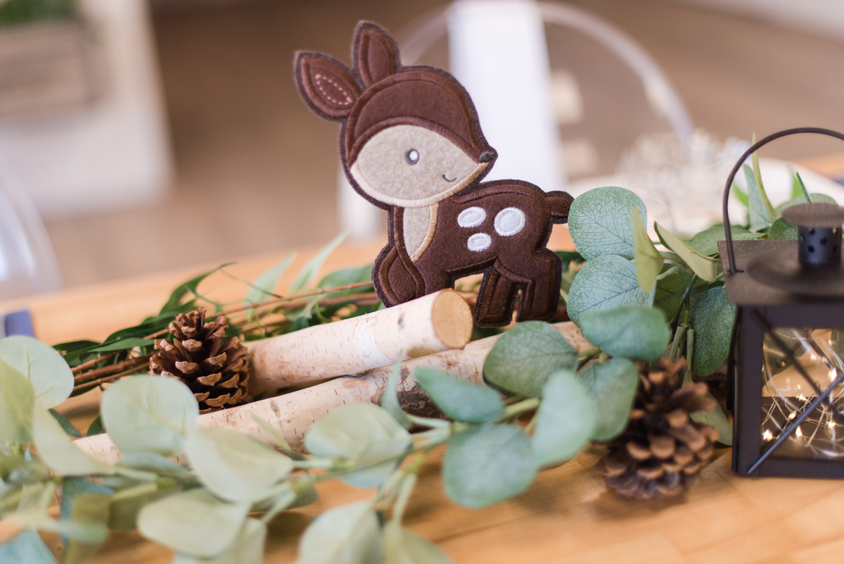 Woodland-themed baby boy shower table centerpiece featuring rustic wood logs, lush greenery, a decorative lantern, and a fabric deer.