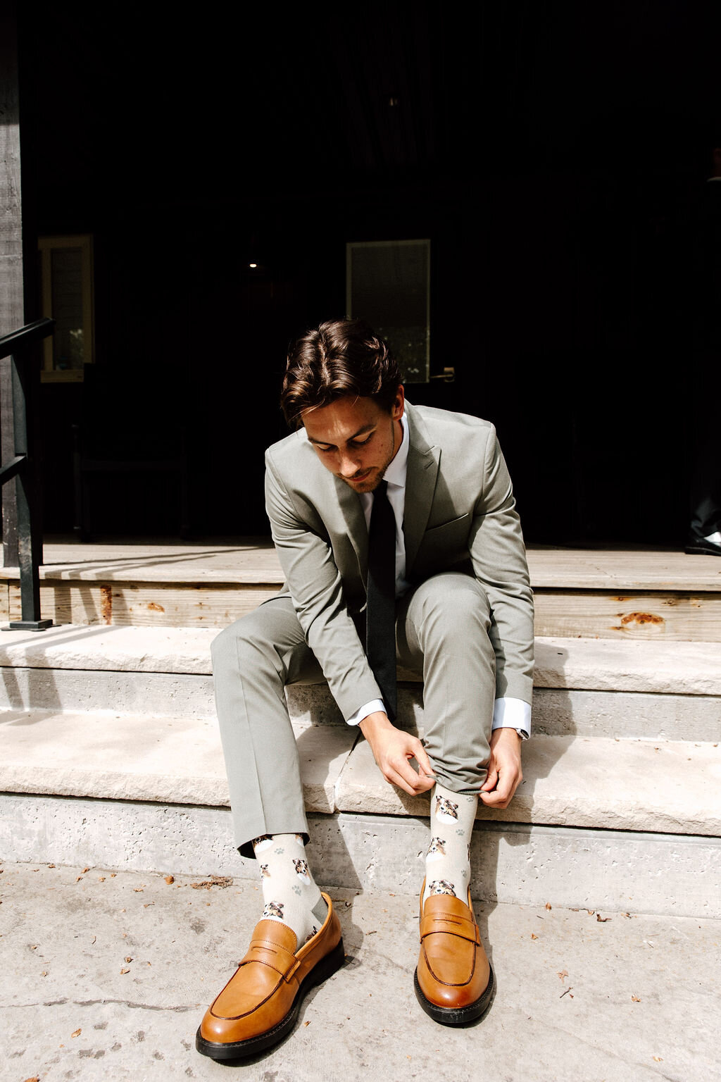 groom in a sage green suit putting his patterned socks on before the wedding in the preparation cottage on the grounds of Willowbrook wedding venue