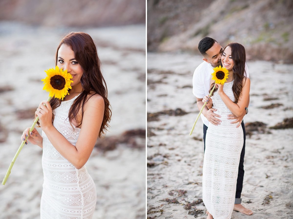Bride to be smiles at the camera while sniffing a sunflower and then her fiance embraces her from behind as they stand on the beach