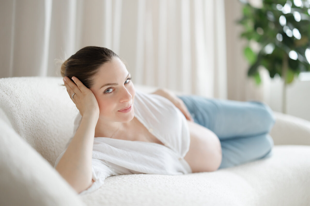 a pregnant women lays on her side on a white fuzzy cough as her white button up shirt parts for her baby bump.