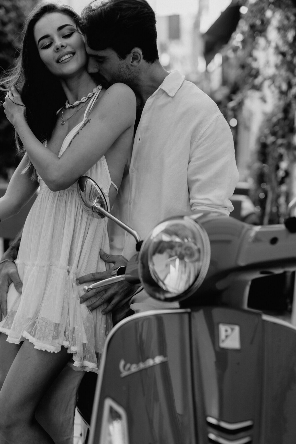 Black and white photo of couple leaning on a scooter while she leans her back against him and he kisses her neck.