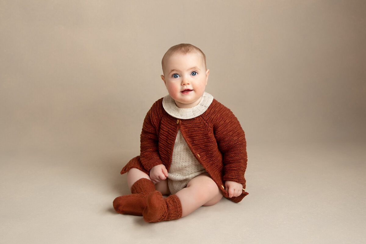 Six month old baby girl dressed in a brown cardigan and socks on a simple cream background.