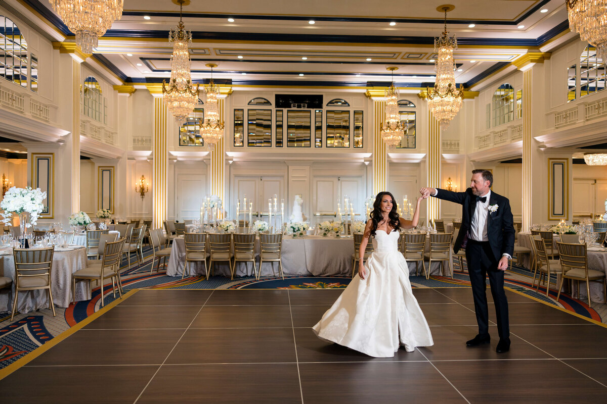 A bride and groom dance joyfully on a delicate, dark-colored floor in an opulent ballroom adorned with crystal chandeliers and elegantly set tables. The room exudes a luxurious wedding atmosphere.
