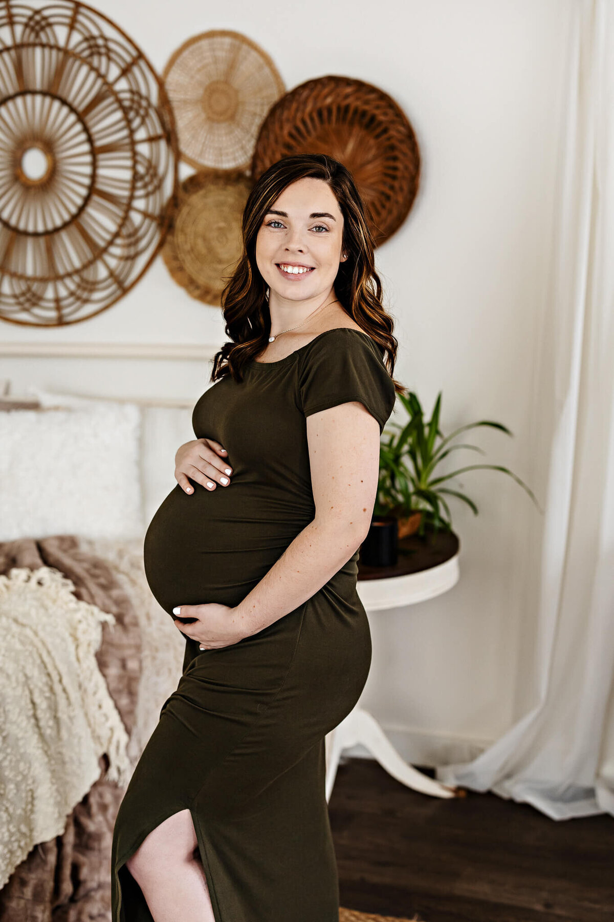 A pregnant woman in a dark green dress stands indoors, smiling with one hand on her belly—a perfect moment for maternity portraits. Behind her are decorative woven wall hangings, a plant on a table, and a cozy blanket on a bed.