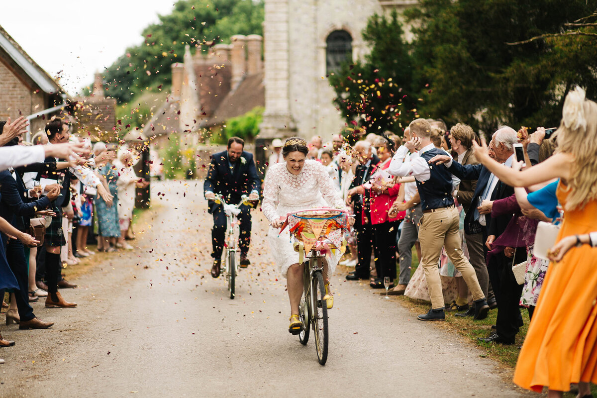 Bride and groom riding bicycles through a cloud of confetti