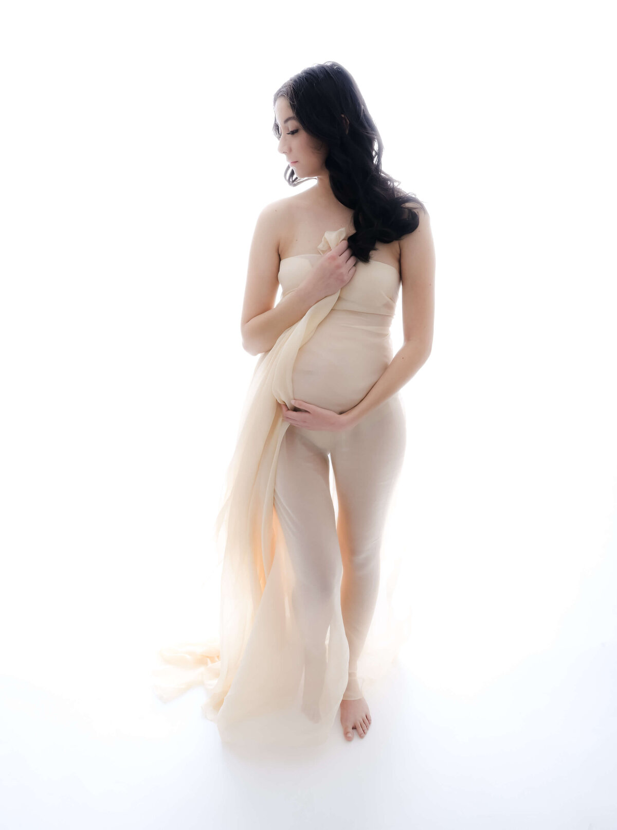 Backlit maternity photo session on a white background where expecting mother is wrapped in an off white sheer fabric and glances over her shoulder as she cradles her growing belly. Image captured by E. Benecki Photogrpahy