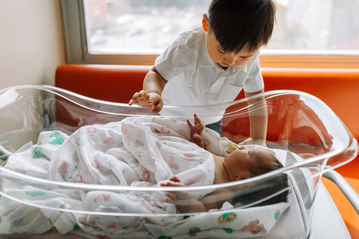 toddler standing on couch to look into hospital bassinet to see newborn sister