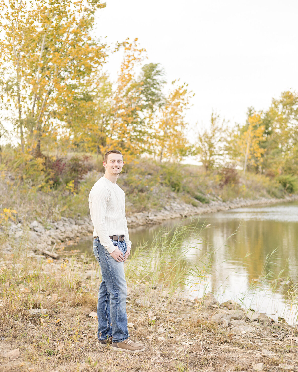 high school senior male, standing next to a pond, with trees in the background, thumbs in his pockets, wearing a light tan long sleeve shirt, blue jeans, a belt, and tan dress shoes, turned to the side looking at the camera smiling