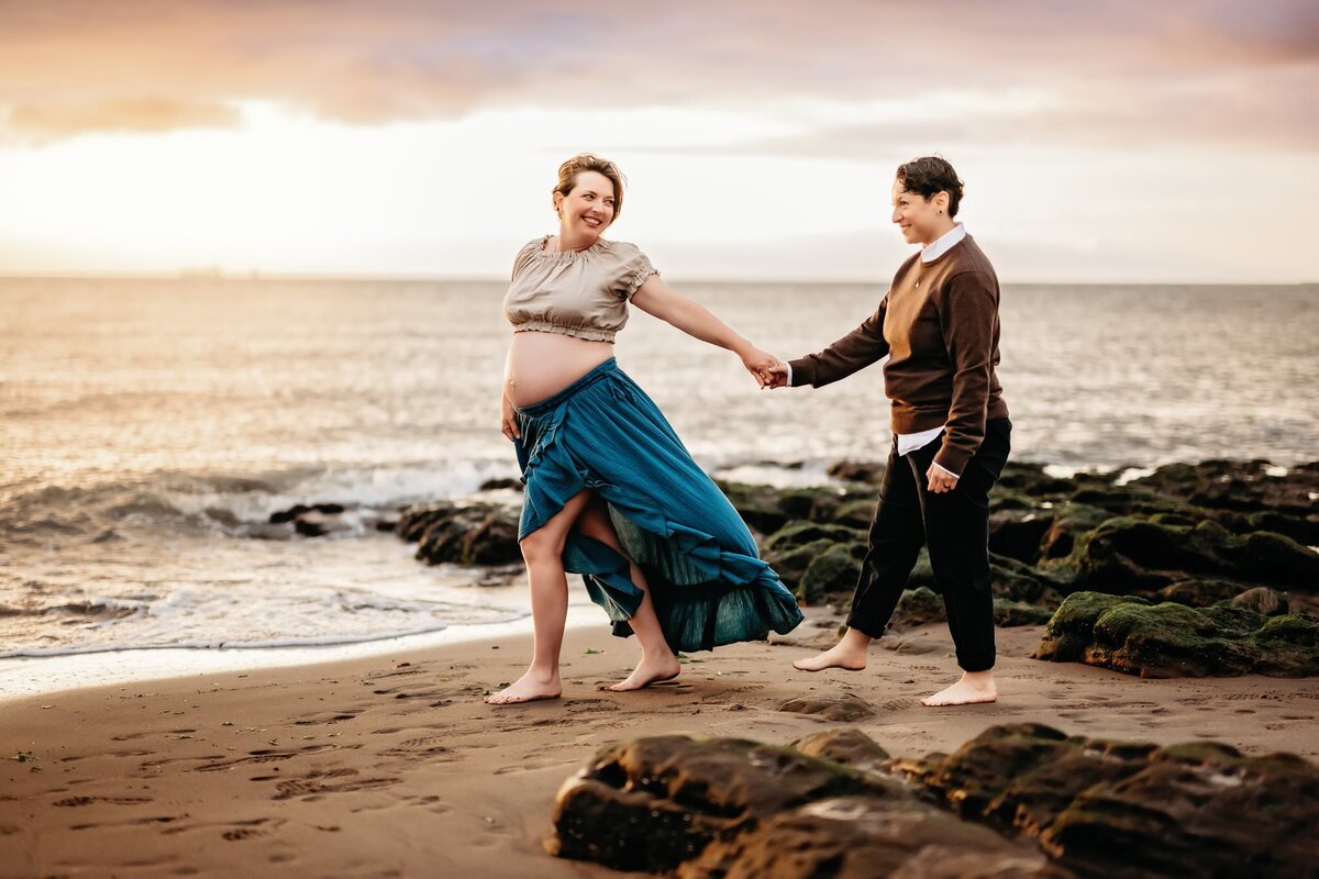 2 mamas at a dreamy beach for their maternity photoshoot. pregnant mama guides her wife by the hand.