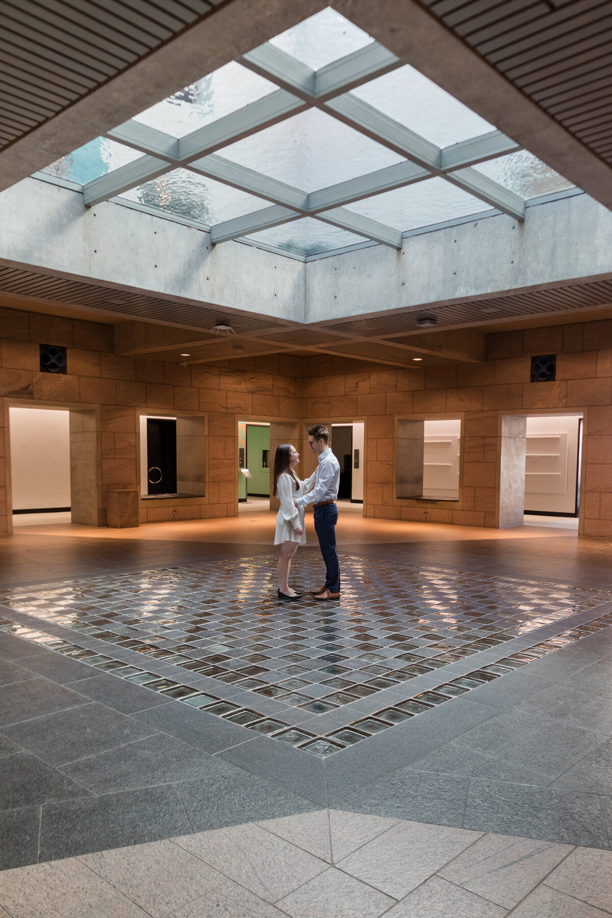 Melissa and Yannick stand beneath the mesmerizing ceiling of the National Gallery, surrounded by the reflective water mirroring their love, creating a magical scene