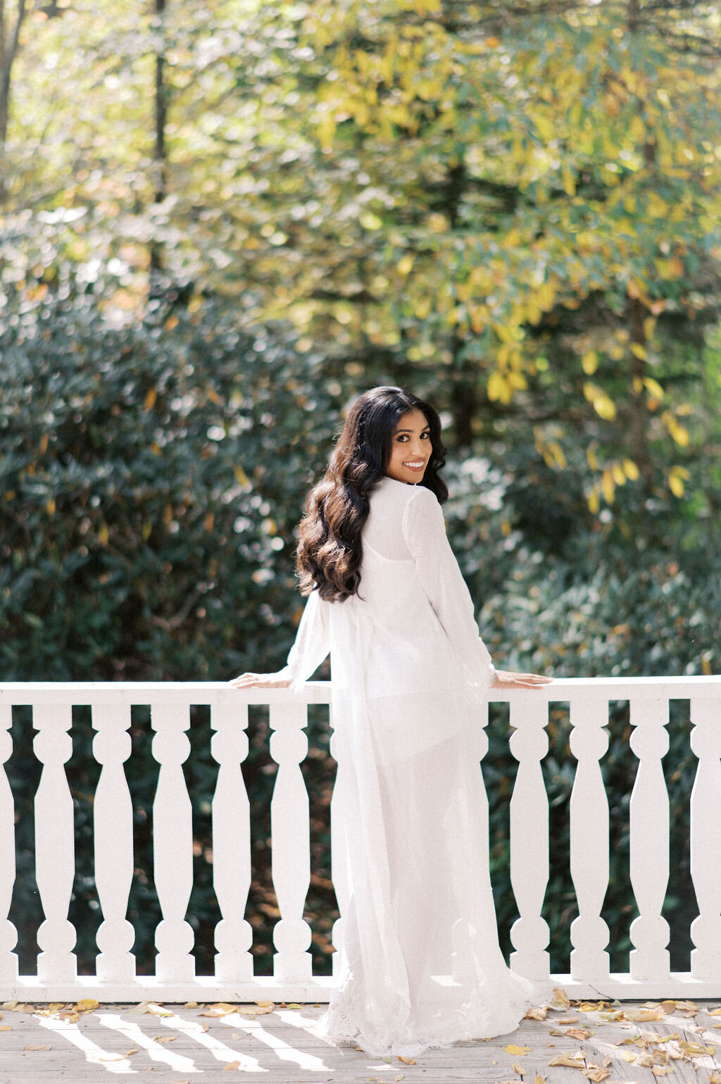 Bride wearing a sheer white robe smiling over her shoulder on a balcony with fall foliage behind her.