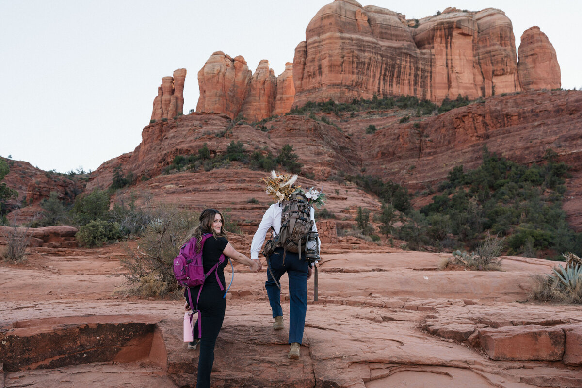 Couple hiking up to their Cathedral Rock elopement in Sedona, Arizona, carrying floral arrangements and backpacks, showcasing a scenic adventure wedding in the red rocks.