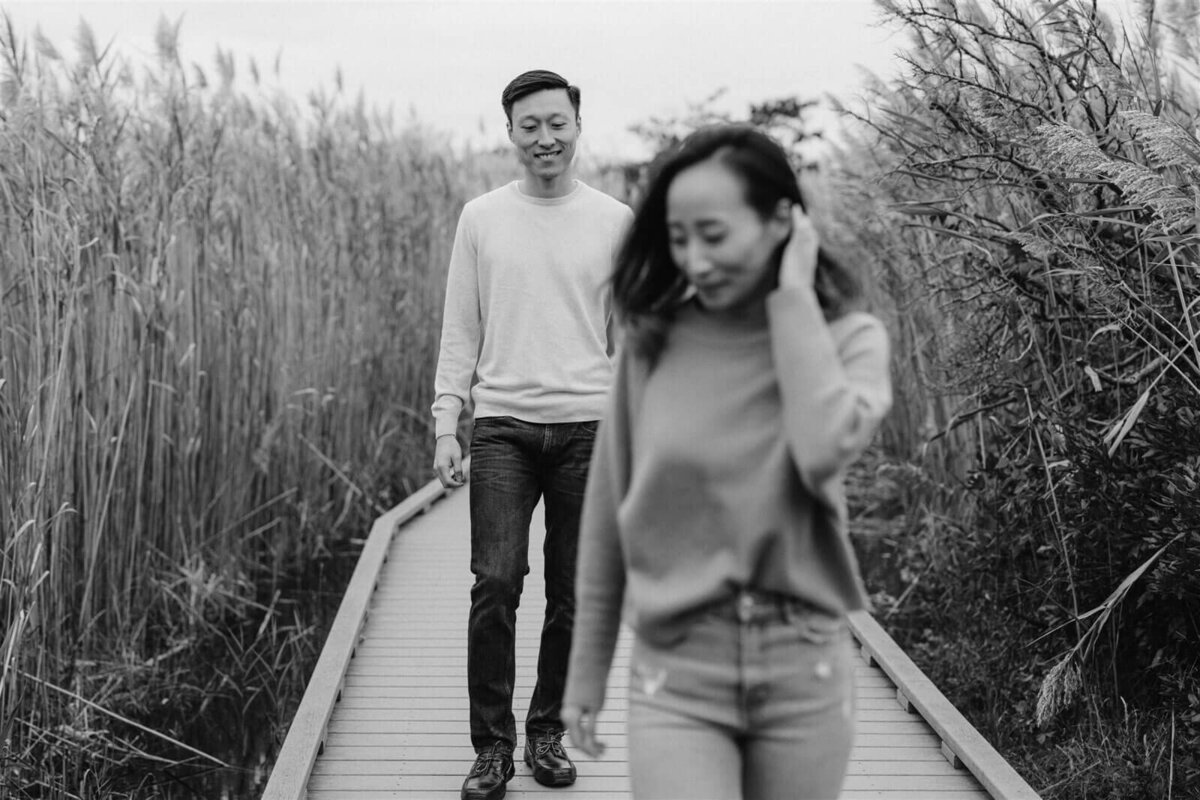 The engaged couple is happily walking on a wooden bridge amidst grasses in New York. Engagement Image by Jenny Fu Studio