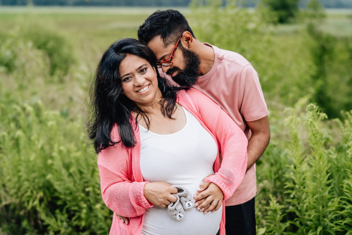 A pregnant woman holds up two little newborn slippers with her husband hugging her from behind.