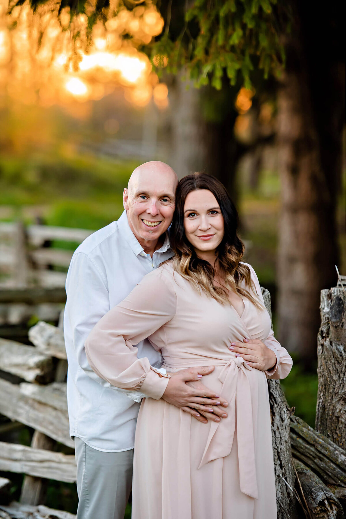 husband and wife standing near a wood fence hugging from behind cradling mom's belly for their Toronto maternity photos.